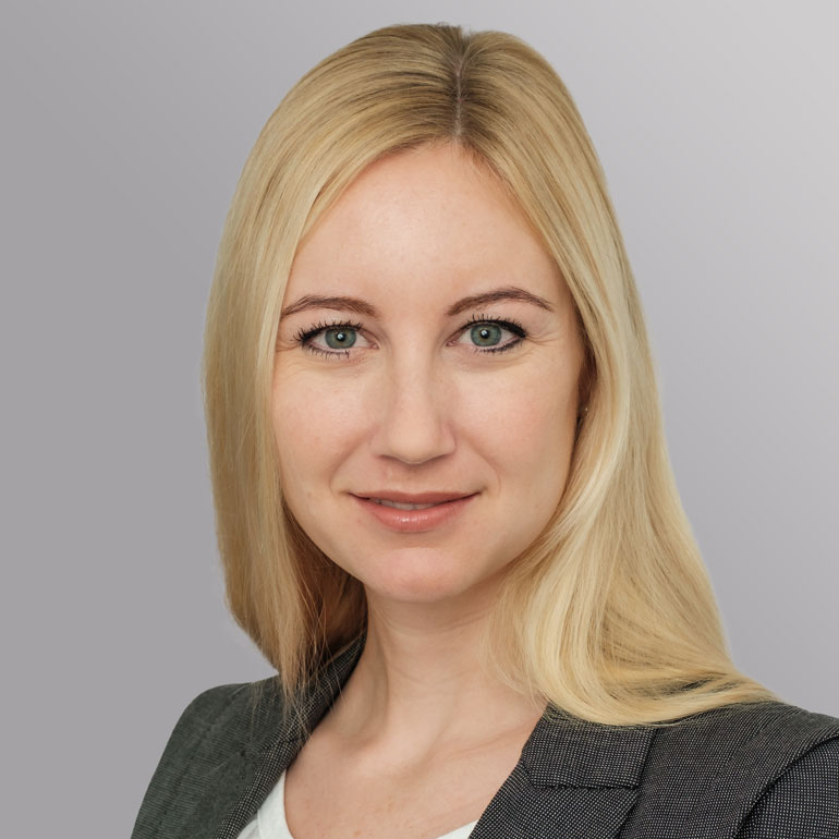 Porträt einer leicht lächelnden Frau mit langen, blonden Haaren und grünen Augen, die einen grauen Blazer trägt. Der Hintergrund ist in sanften Grautönen gehalten.