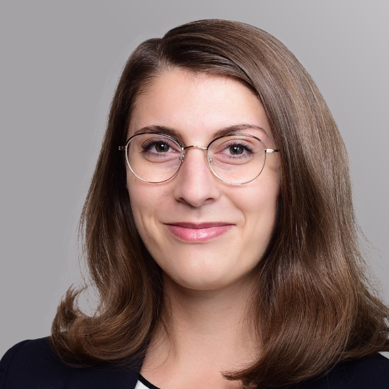 Ein Porträt einer jungen Frau mit langen, lockigen, braunen Haaren. Sie trägt eine schwarze Brille und hat ein freundliches Lächeln. Die Hintergrundfarbe ist grau und sorgt für einen neutralen Kontrast zu ihrem Gesicht.