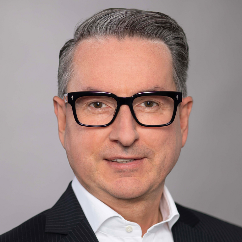 Portrait eines Mannes mit grauen Haaren, Brille und einem freundlichen Ausdruck. Er trägt ein schwarzes Jackett über einem weißen Hemd. Der Hintergrund ist schlicht und hell.