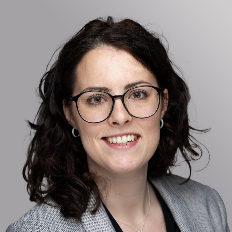 Porträt einer lächelnden Frau mit lockigem, dunklem Haar, die eine Brille trägt. Sie hat eine graue Blazerjacke an und präsentiert ein freundliches, professionelles Auftreten vor einem hellen, einfarbigen Hintergrund.