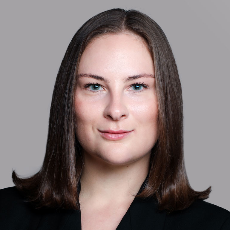 Porträt einer Frau mit langen, glatten Haaren und blauen Augen. Sie trägt einen schwarzen Blazer und hat einen neutralen Gesichtsausdruck. Der Hintergrund ist grau.