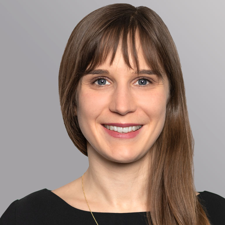 Lächelnde Frau mit langen, braunen Haaren, die an einem hellen Hintergrund steht. Sie trägt ein schwarzes Oberteil und eine feine Halskette. Ihre Augen sind blau, und sie vermittelt einen freundlichen und offenen Eindruck.