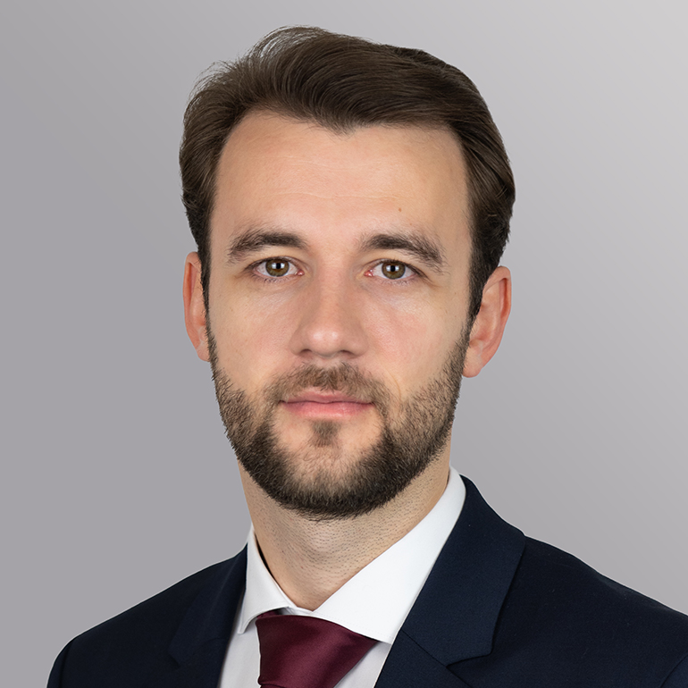 Porträt eines Mannes mit kurz geschnittenem Haar und Bart. Er trägt einen dunkelblauen Anzug und eine rote Krawatte. Der Hintergrund ist grau. Der Mann schaut neutral und freundlich zur Kamera.