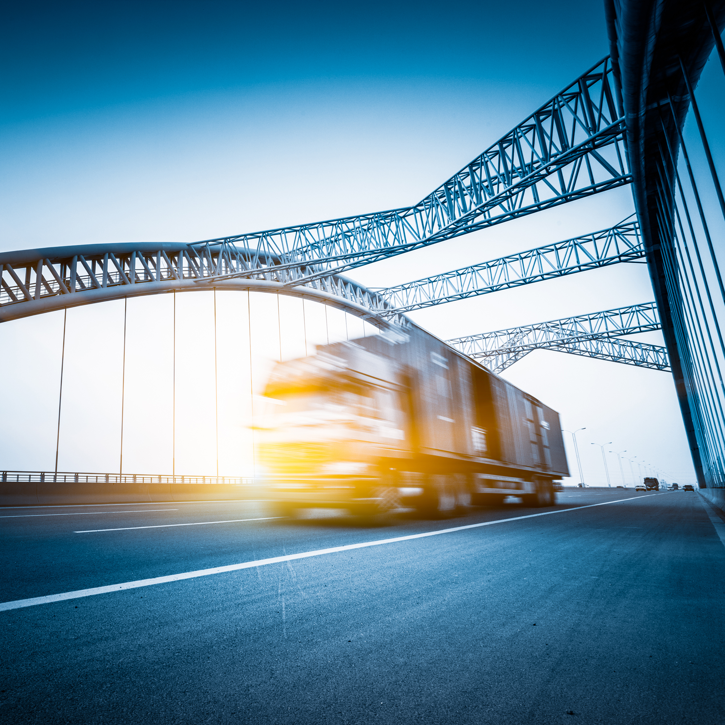 A blurred truck is shown in motion on a highway, passing beneath a large, modern bridge with intricate metal design. The scene conveys a sense of speed and transportation against a light blue sky.
