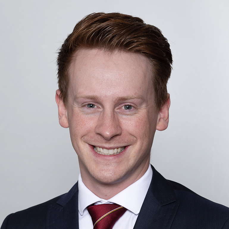 Portrait eines lächelnden Mannes mit kurzen, roten Haaren, der einen dunklen Anzug mit einer roten Krawatte trägt. Der Hintergrund ist hell und neutral.