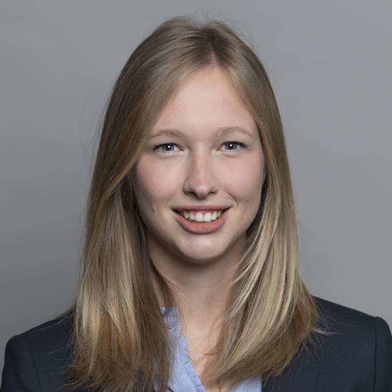 Blonde Frau mit langen Haaren lächelt direkt in die Kamera. Sie trägt einen dunkelblauen Blazer und ein hellblaues Hemd. Der Hintergrund ist grau. Ihr Ausdruck vermittelt Freundlichkeit und Zuversicht.