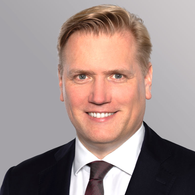 Ein lächelnder Mann mit blonden Haaren trägt einen dunklen Anzug und eine hellblaue Krawatte. Er hat eine freundliche Ausstrahlung und schaut direkt in die Kamera. Der Hintergrund ist grau und neutral gehalten.