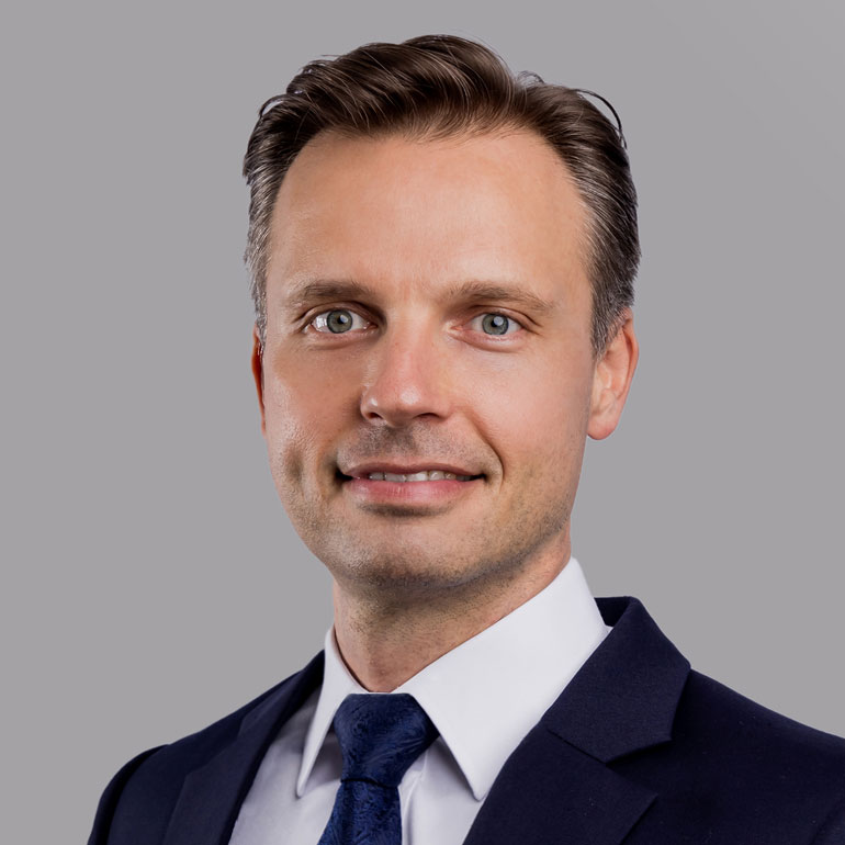 Porträt eines lächelnden Mannes mit kurzem, gestyltem Haar. Er trägt einen dunkelblauen Anzug und eine hellblaue Krawatte. Der Hintergrund ist grau. Der Mann hat blaue Augen und wirkt freundlich und professionell.