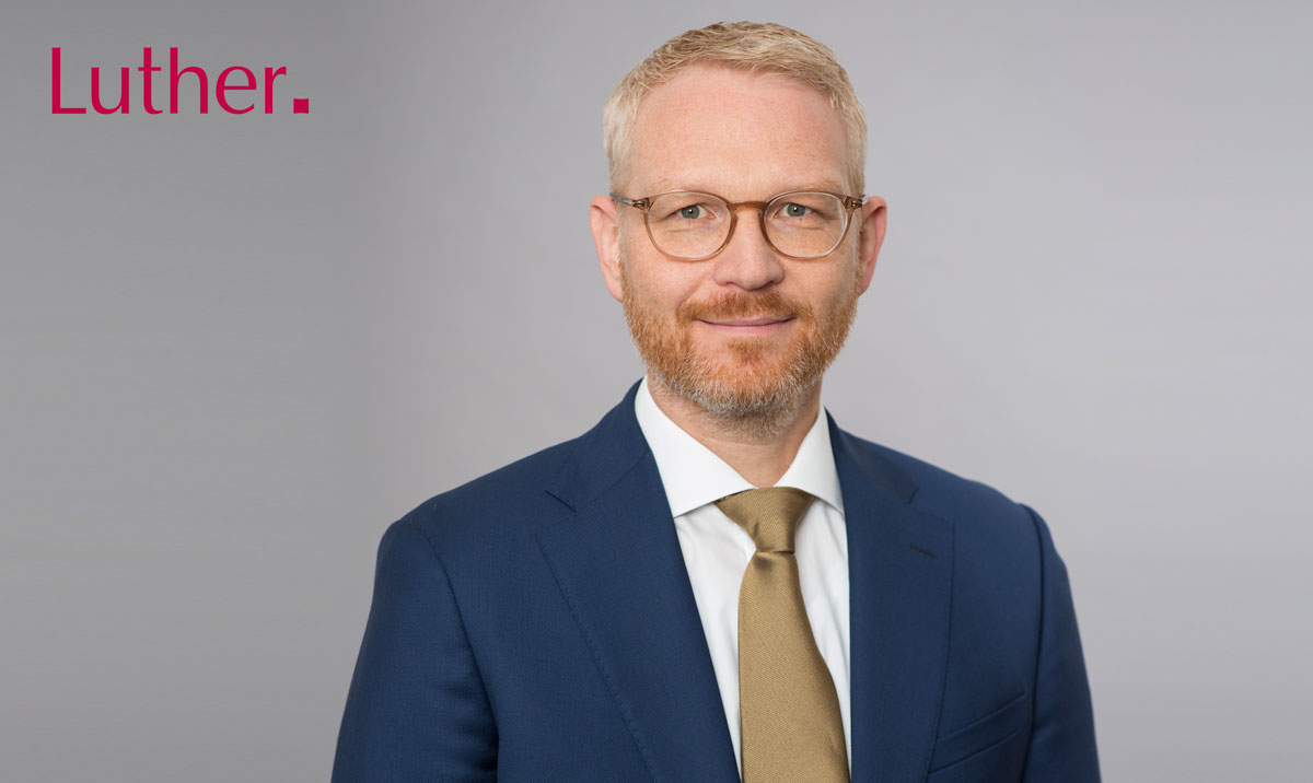 A professional man with a light beard and glasses smiles at the camera. He is wearing a dark blue suit and a light brown tie. The background is light grey, and the word "Luther." is displayed prominently in pink on the left side.