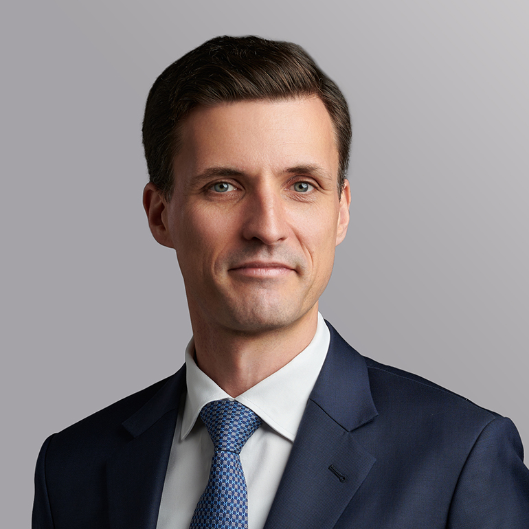 Porträt eines Mannes mit kurzer, dunkler Frisur, der in einem dunkelblauen Anzug und einer hellblauen Krawatte gekleidet ist. Er schaut leicht lächelnd in die Kamera, vor einem neutralen grauen Hintergrund.