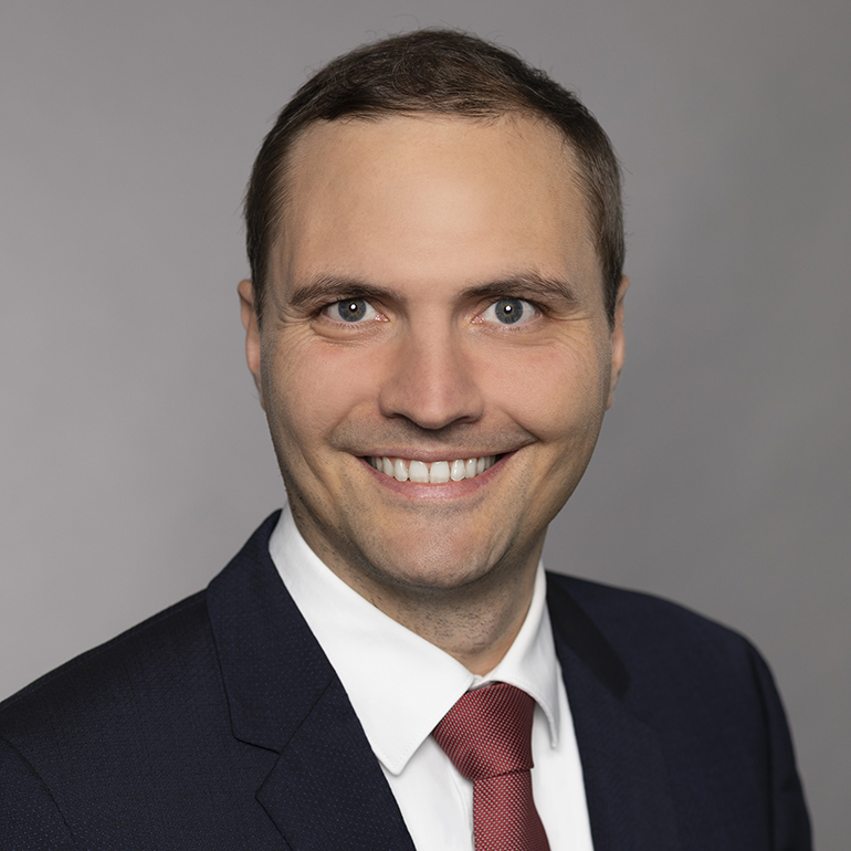 Ein lächelnder Mann mit kurzen Haaren trägt einen dunklen Anzug und eine weiße Hemd mit roter Krawatte. Der Hintergrund ist neutral grau. Sein freundlicher Blick vermittelt Selbstbewusstsein.