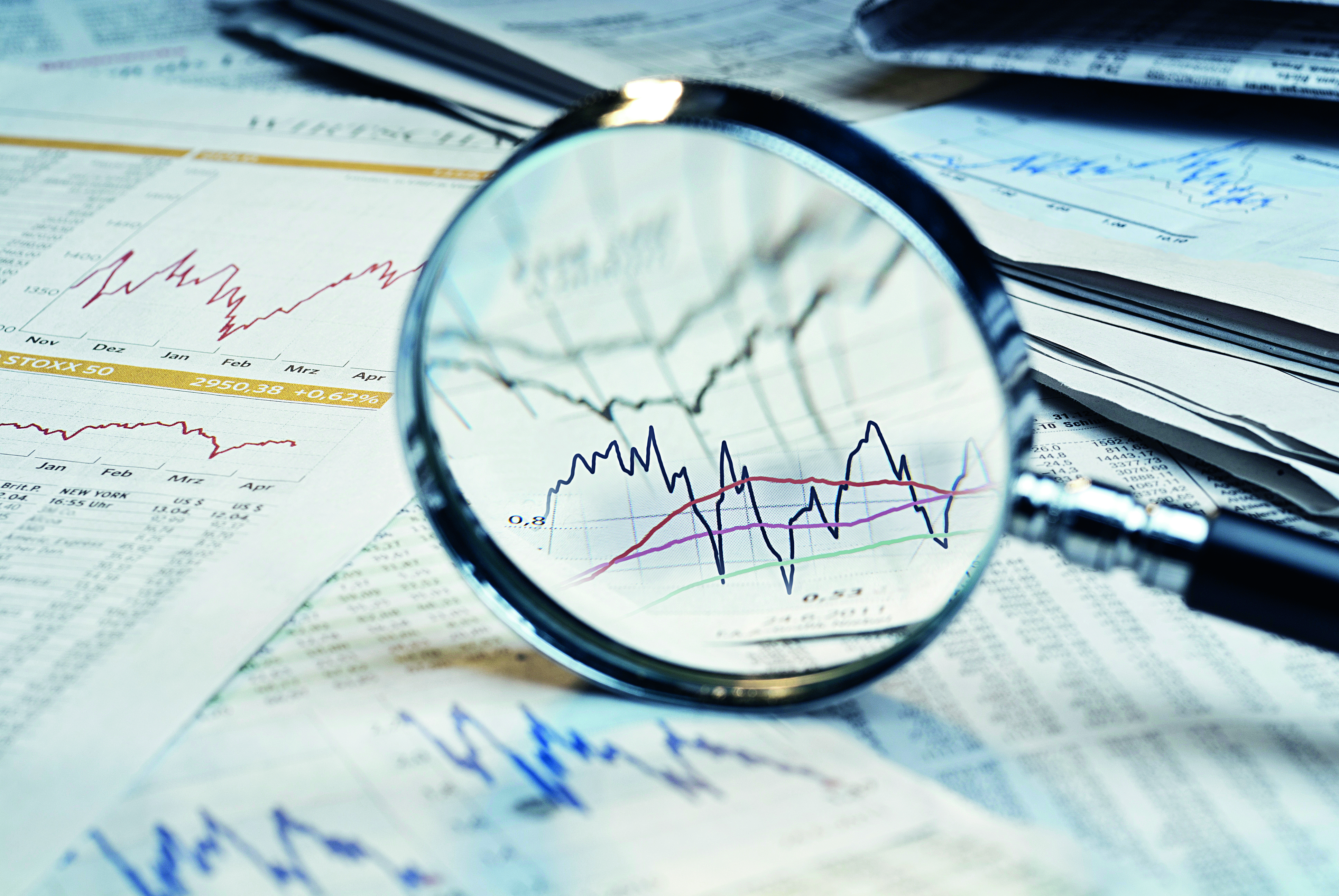 Eine Lupe fokussiert Diagramme und Tabellen mit Finanzdaten und Kursentwicklungen. Die Diagramme zeigen verschiedene Kursverläufe, die durch Linien graphisch dargestellt sind. Der Hintergrund besteht aus weiteren Finanzunterlagen.