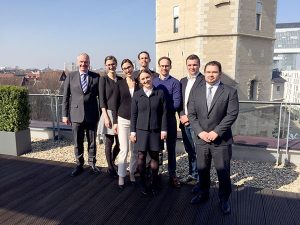Eine Gruppe von sieben Personen steht auf einer Terrasse. Sie tragen formelle Kleidung und lächeln in die Kamera. Im Hintergrund ist ein Gebäude sichtbar. Die Szene vermittelt eine freundliche, professionelle Atmosphäre.