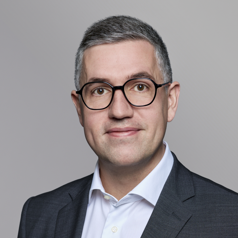 Ein lächelnder Mann mit Brille und kurzen grauen Haaren trägt ein dunkelgraues Sakko über einem weißen Hemd. Der Hintergrund ist neutral und einfarbig. Der Ausdruck des Mannes ist freundlich und zugewandt.