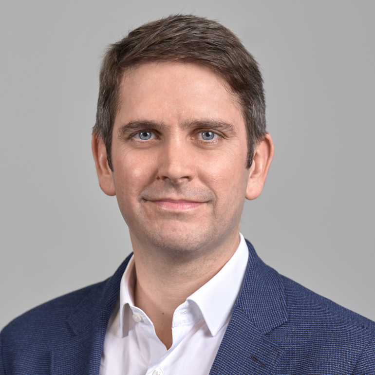 Porträt eines Mannes mit kurzem, braunem Haar und blauen Augen. Er trägt ein weißes Hemd und einen blauen Blazer. Der Hintergrund ist grau. Der Mann hat einen freundlichen Gesichtsausdruck und schaut direkt in die Kamera.