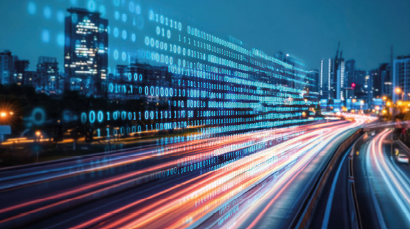 Ein Baustellenfahrzeug, ein Straßenwalze, ist auf einer neu asphaltierten Straße bei Sonnenuntergang unterwegs. Im Hintergrund sind Bäume und weitere Fahrzeuge sichtbar. Die warme Farben des Himmels vermitteln eine ruhige Stimmung.