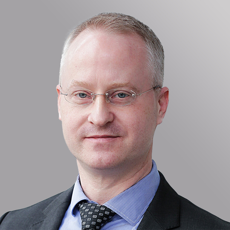 Ein Mann mit kurzen, grauen Haaren und einer Brille trägt einen schwarzen Anzug und ein gestreiftes Hemd. Er schaut direkt in die Kamera und hat einen neutralen Gesichtsausdruck. Der Hintergrund ist einfarbig grau.