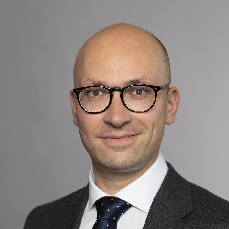 Ein Mann mit Glatze und Brille lächelt in die Kamera. Er trägt ein weißes Hemd mit einer dunklen Krawatte mit Punkten und einen grauen Anzug. Der Hintergrund ist einfarbig grau.