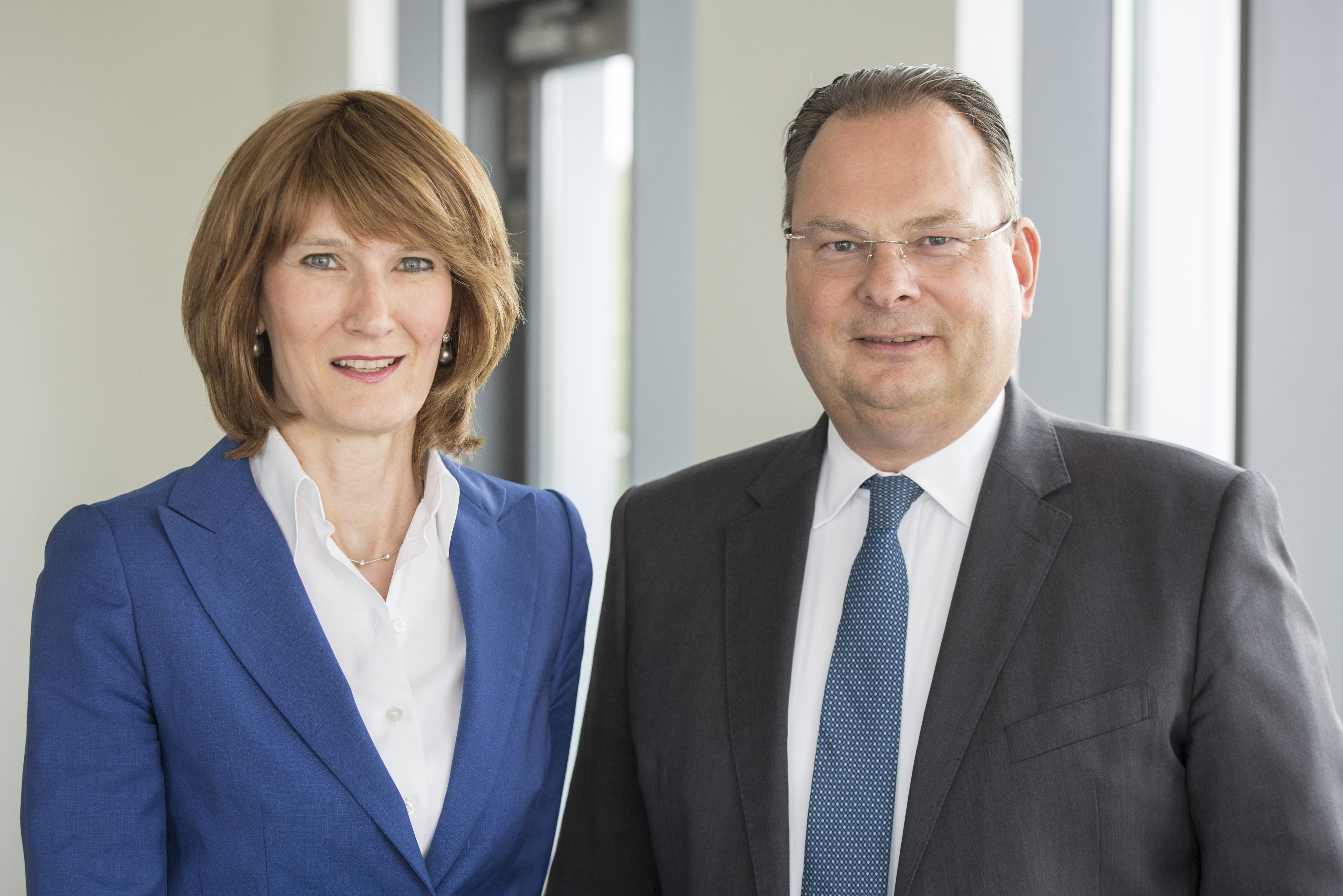 Zwei Personen stehen in einem modernen Bürointerieur. Die Frau trägt einen blauen Blazer und hat schulterlange braune Haare. Der Mann ist in einen dunklen Anzug gekleidet und trägt eine hellblaue Krawatte. Beide blicken freundlich in die Kamera.