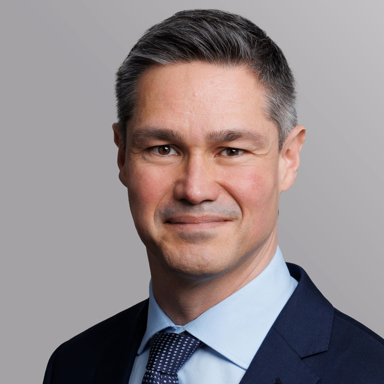 Porträt eines Mannes mit kurzen, grau melierten Haaren und einem freundlichen Lächeln. Er trägt ein dunkelblaues Anzugsjackett und ein hellblaues Hemd mit einer gemusterten Krawatte. Der Hintergrund ist sanft grau.