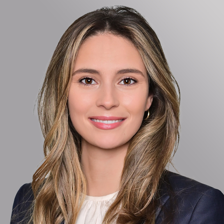 Porträt einer jungen Frau mit langen, welligen Haaren. Sie trägt ein dunkles Blazer-ähnliches Outfit und hat ein freundliches Lächeln. Der Hintergrund ist grau. Ihre Ausstrahlung wirkt professionell und einladend.