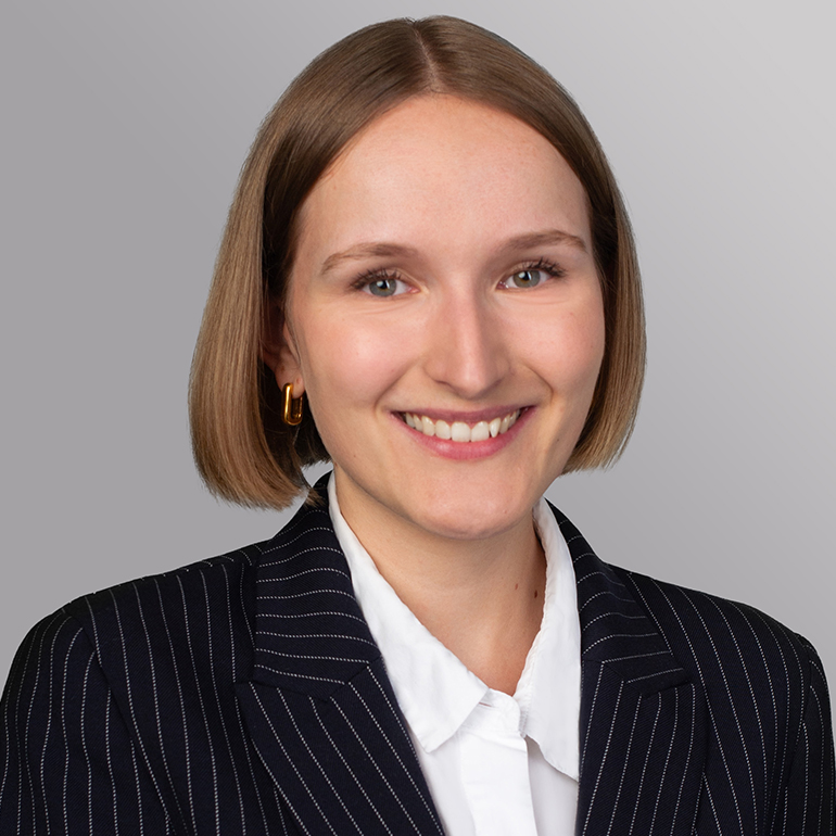 Ein Porträt einer jungen Frau mit kurzen, blonden Haaren und einem freundlichen Lächeln. Sie trägt einen dunkel gestreiften Blazer über einem weißen Hemd. Der Hintergrund ist grau und schlicht, wodurch die Frau im Vordergrund hervortritt.