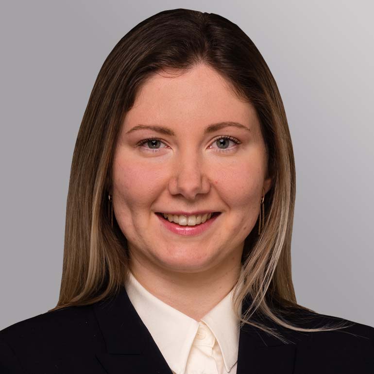 Ein Porträt einer lächelnden Frau mit langen, hellen Haaren. Sie trägt ein schwarzes Jackett und ein helles Hemd. Der Hintergrund ist grau. Ihr Ausdruck wirkt freundlich und professionell.