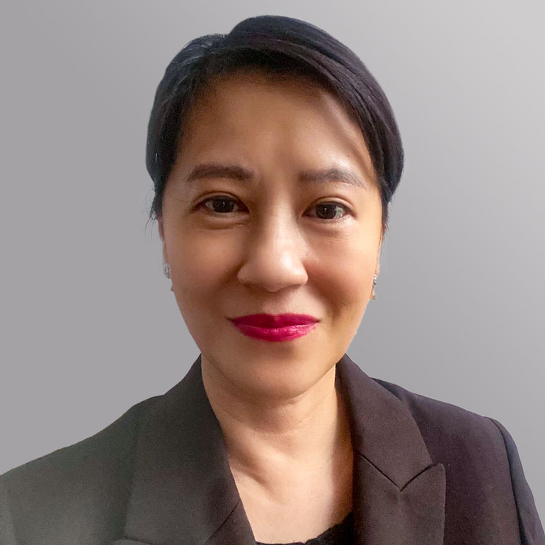 Porträt einer Frau mit kurzem, dunklem Haar, die einen blauen Blazer trägt. Sie hat einen freundlichen Ausdruck und trägt rote Lippenfarbe. Der Hintergrund ist grau.