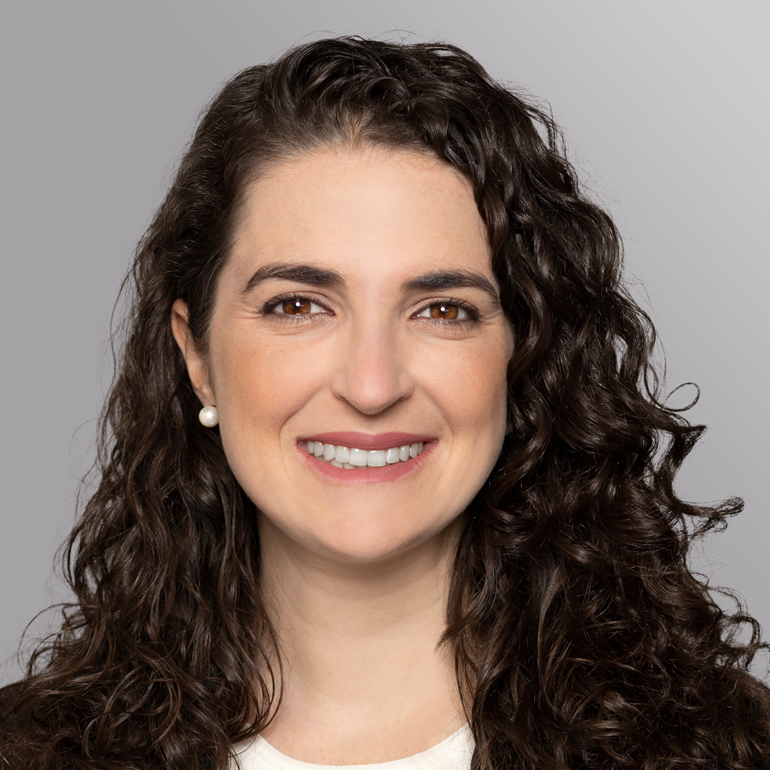 Porträt einer Frau mit lockigem, braunem Haar, die lächelt. Sie trägt ein helles Oberteil und Perlenohrringe. Der Hintergrund ist neutral und grau, was den Fokus auf ihr Gesicht lenkt.