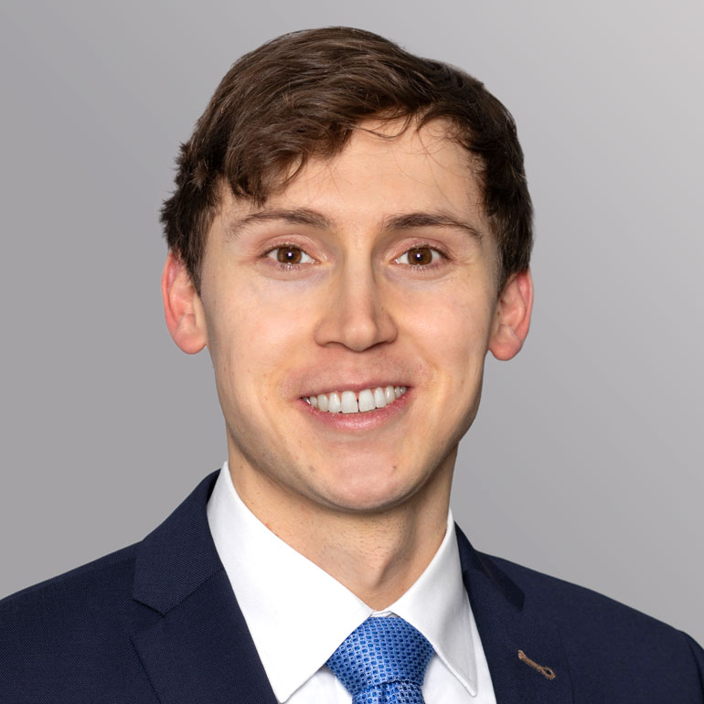 Eine lächelnde Person mit kurzem, braunem Haar trägt einen dunkelblauen Anzug und eine hellblaue Krawatte. Der Hintergrund ist grau. Die Person hat einen freundlichen Ausdruck und zeigt Zähne.