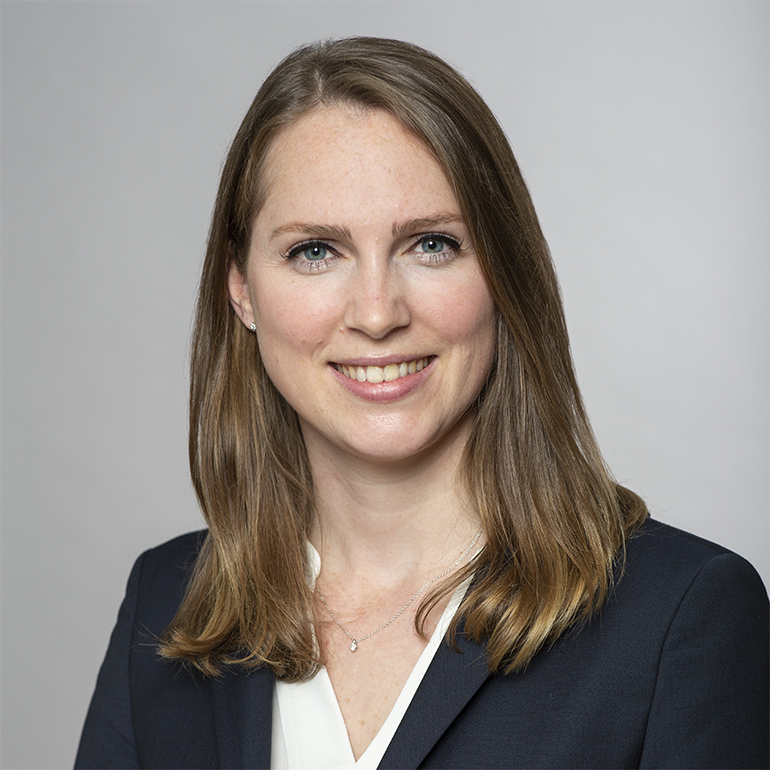 Porträt einer lächelnden Frau mit langen, glatten, braunen Haaren. Sie trägt ein dunkles Blazer-Oberteil und ein helles Oberteil darunter. Der Hintergrund ist neutral und verschwommen, was den Fokus auf ihr Gesicht lenkt.