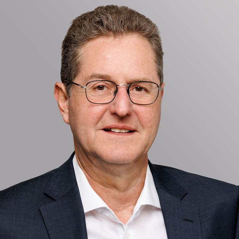 Porträt eines Mannes mit kurzem, grauen Haar, der eine Brille trägt. Er hat ein freundliches Lächeln und trägt einen dunklen Anzug mit einem weißen Hemd. Der Hintergrund ist grau.