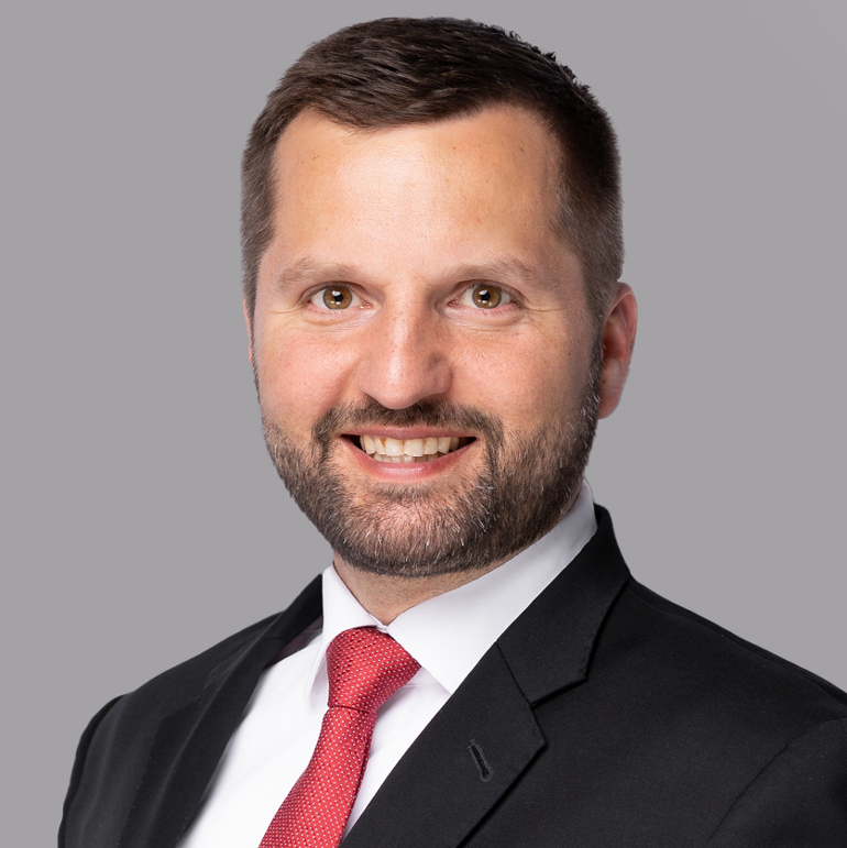 Porträt eines Mannes mit kurzem Haar und einem gepflegten Bart. Er trägt einen schwarzen Anzug mit weißem Hemd und roten Krawatte. Der Mann lächelt freundlich in die Kamera, vor einem grauen Hintergrund.