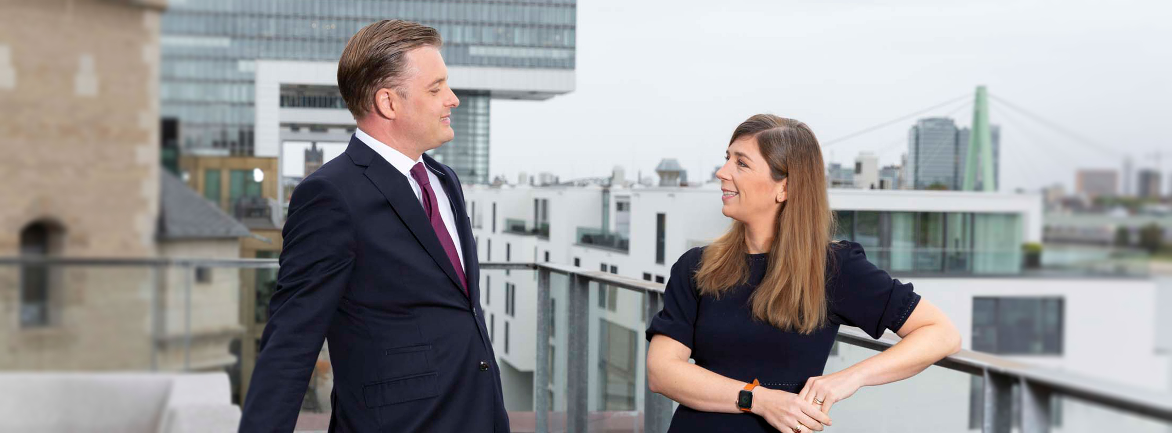 Ein Mann und eine Frau stehen auf einer Terrasse und schauen sich an. Beide tragen formelle Kleidung. Im Hintergrund sind moderne Gebäude und eine Brücke sichtbar. Die Atmosphäre wirkt freundlich und geschäftlich.