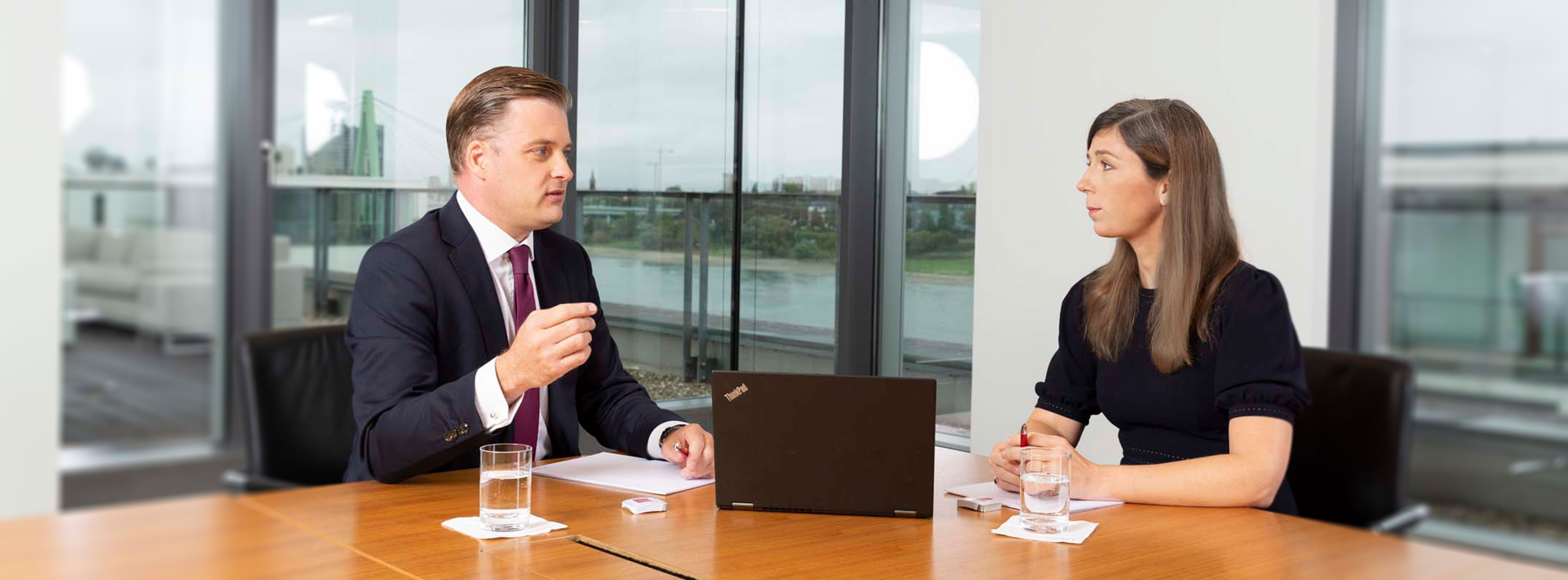 Zwei Personen führen ein Gespräch in einem Konferenzraum. Ein Mann trägt einen Anzug und spricht gestikulierend, während eine Frau sitzt und ihm aufmerksam zuhört. Vor ihnen befindet sich ein Laptop und Gläser mit Wasser auf dem Tisch.