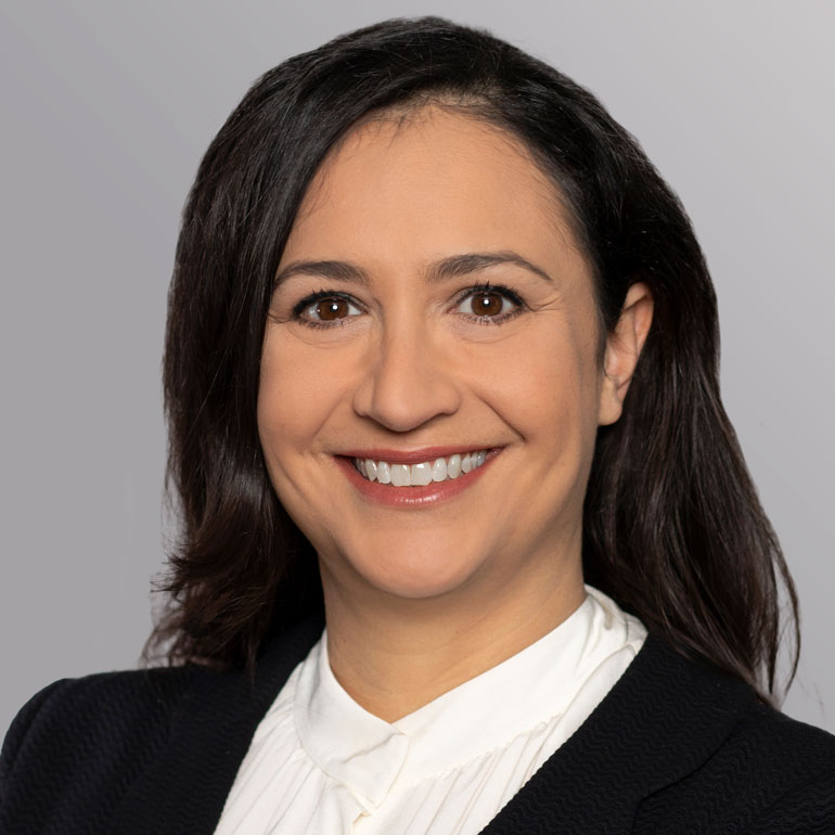 Porträt einer lächelnden Frau mit dunklen Haaren, die in einem weißen Oberteil und einem schwarzen Blazer gekleidet ist. Der Hintergrund ist grau und unauffällig, um den Fokus auf das Gesicht zu lenken.