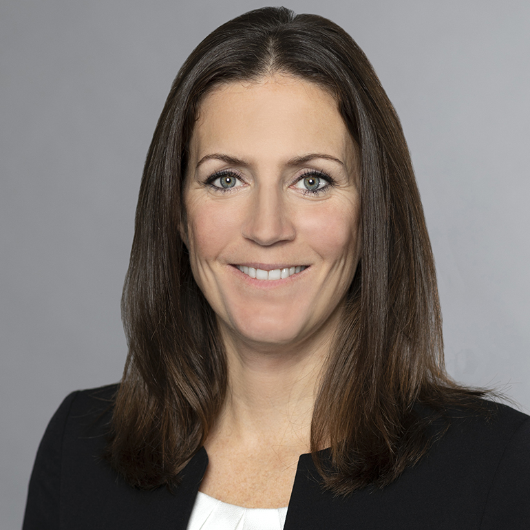 Porträt einer lächelnden Frau mit langen, braunen Haaren. Sie trägt einen dunklen Blazer und hat einen neutralen Hintergrund. Ihr Gesicht zeigt eine freundliche und professionelle Ausstrahlung.