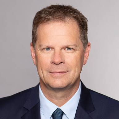 Porträt eines Mannes mit kurzen, braunen Haaren und einem neutralen Gesichtsausdruck. Er trägt einen dunklen Anzug und ein hellblaues Hemd. Der Hintergrund ist schlicht und hell, wodurch der Fokus auf dem Gesicht liegt.