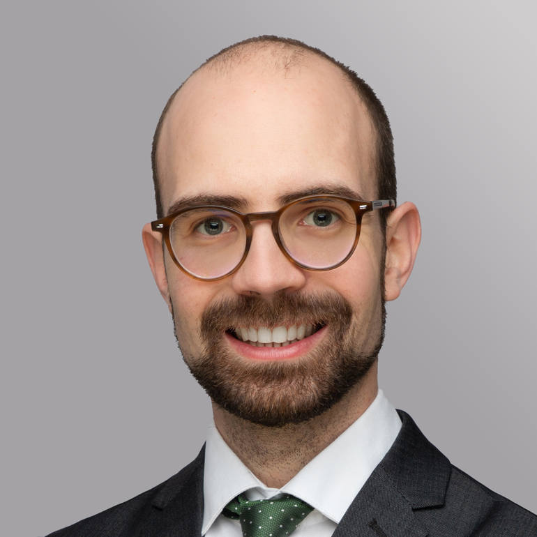 Ein junger Mann mit Brille lächelt freundlich. Er trägt einen Anzug mit einer weißen Hemd und einer grünen Krawatte mit Punkten. Der Hintergrund ist einfarbig und grau.