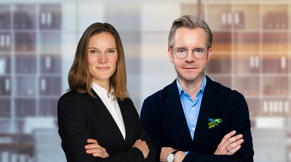 A professional woman in a black blazer and a man in a navy jacket stand confidently with their arms crossed. They are set against a blurred, modern office background, conveying a sense of teamwork and professionalism.