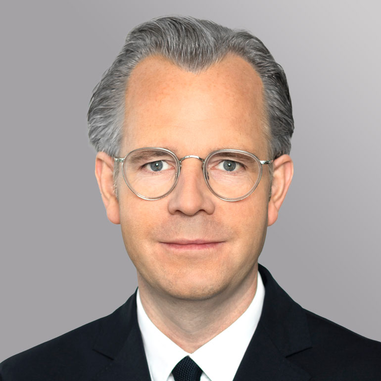 Porträt eines Mannes mit grauen Haaren und Brille. Er trägt einen schwarzen Anzug mit Krawatte und schaut ernst in die Kamera. Der Hintergrund ist grau und neutral gehalten.