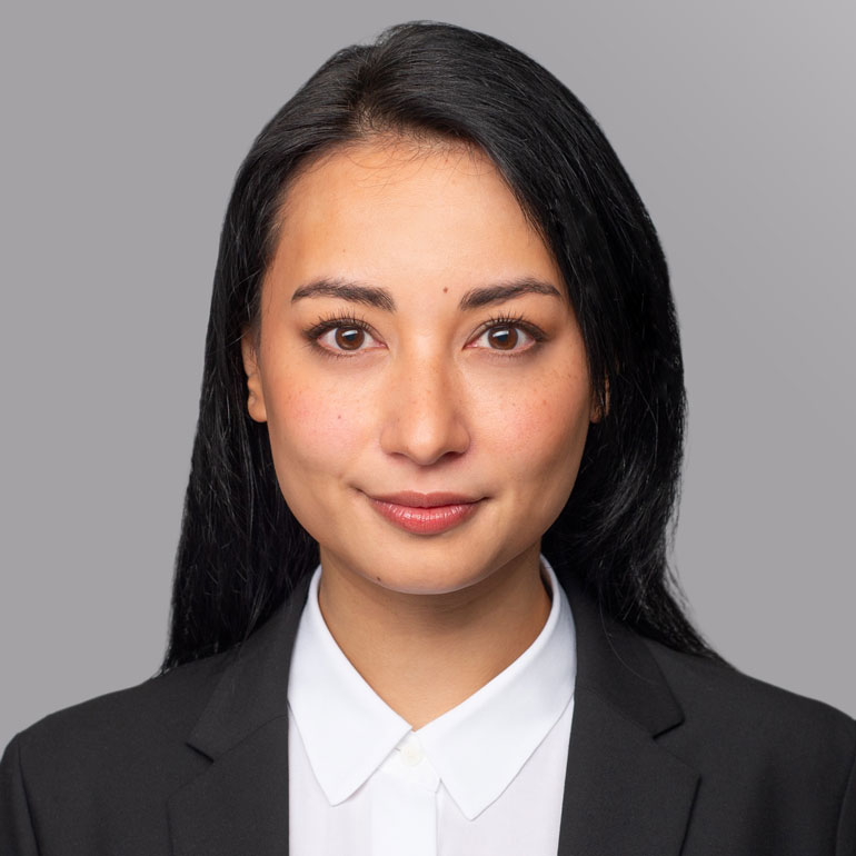 Ein Portrait einer Frau mit langen, schwarzen Haaren, die ein schwarzes Sakko und ein weißes Hemd trägt. Sie blickt direkt in die Kamera mit neutralem Gesichtsausdruck vor einem grauen Hintergrund.