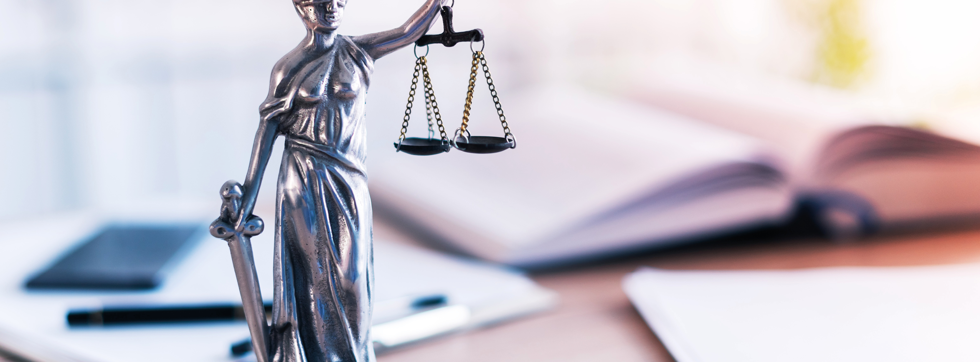 Eine Statue von Justitia, die mit einer Waage in der linken Hand und einem Schwert in der rechten Hand dargestellt ist. Der Hintergrund zeigt verschwommene Bücher und Schreibmaterialien, was auf rechtliche Themen hinweist. Die Statue symbolisiert Gerechtigkeit und Rechtsprechung.