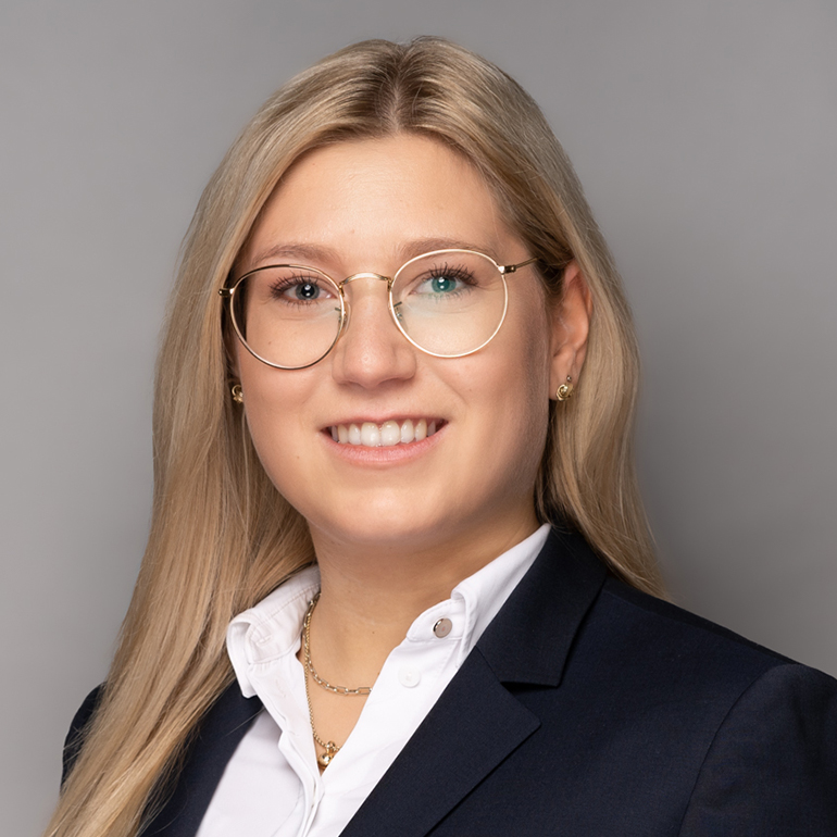 Ein Porträt einer jungen Frau mit langen, blonden Haaren. Sie trägt eine dunkle Blazerjacke und eine weiße Bluse. Die Frau lächelt freundlich und hat eine moderne Brille auf. Der Hintergrund ist grau und schlicht gehalten.