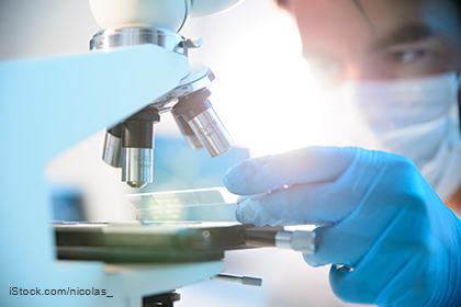 Ein Wissenschaftler in Schutzkleidung arbeitet am Mikroskop. Er hält eine Objektträgerplatte in der Hand und bereitet sie zur Untersuchung vor. Die Umgebung ist hell und die Konzentration des Forschers ist deutlich spürbar.