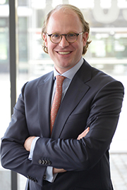 A smiling man stands confidently with arms crossed, wearing glasses and a business suit with a blue blazer and a patterned tie. The background appears to be a modern office setting, suggesting a professional environment.