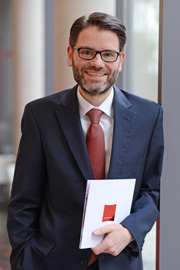 A man with glasses and a beard stands, smiling confidently. He wears a dark suit and a red tie, holding a white folder with a red element on it. The background suggests an indoor setting with natural light.