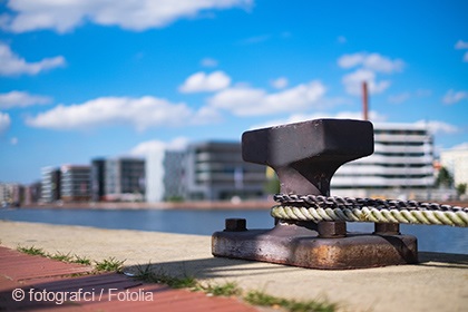 A mooring cleat with a rope is positioned on a waterfront pathway, with modern buildings and a blue sky in the background. The scene conveys a sense of urban harbor life and calm waters.