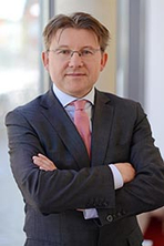 Das Bild zeigt einen Mann mit Brille in einem eleganten Anzug, der mit verschränkten Armen in die Kamera schaut. Der Hintergrund ist hell und modern, was eine professionelle Atmosphäre vermittelt.