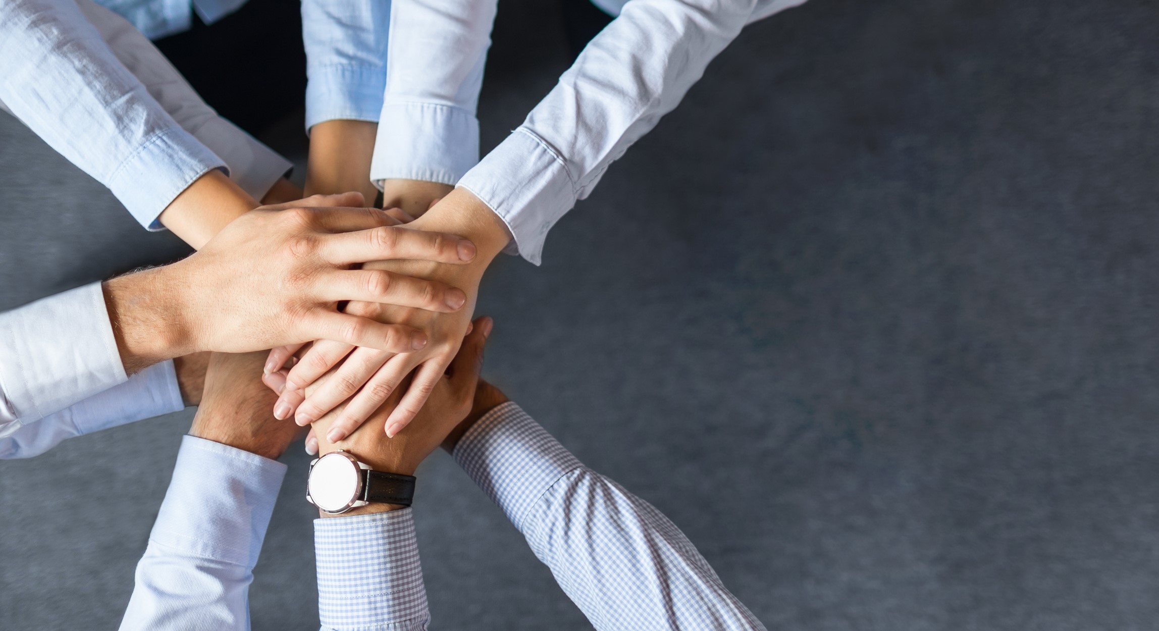 Eine Gruppe von Händen unterschiedlicher Personen liegt übereinander. Die Hände symbolisieren Zusammenarbeit und Teamgeist. Die Menschen scheinen sich zu unterstützen und ein gemeinsames Ziel zu verfolgen. Der Hintergrund ist dunkel und der Fokus liegt auf den Händen.